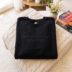May include: A black crewneck sweatshirt featuring the embroidered text "me? overreacting? shit probably." in a matching black thread. The garment has long sleeves, a ribbed neckline, and a standard fit, showcasing a clean and simple design.