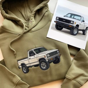 May include: An olive green hooded sweatshirt featuring an embroidered graphic of a white pickup truck. The embroidery depicts the vehicle from a front-side angle with black tyres, a black grille, and orange side marker lights on the front bumper.