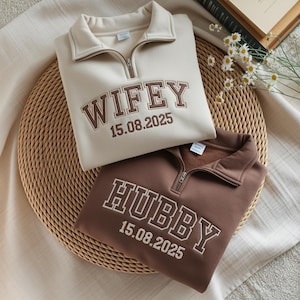 May include: Two quarter-zip sweatshirts, one cream and one brown, are displayed. The cream sweatshirt has "WIFEY 15.08.2025" embroidered in brown. The brown sweatshirt has "HUBBY 15.08.2025" embroidered in white. Both are on a woven mat.