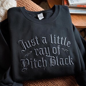 May include: Black crewneck sweatshirt with the embroidered phrase "Just a little ray of Pitch Black" in a gothic font. Two bat silhouettes are embroidered on the design. The sweatshirt is folded on a brown surface.