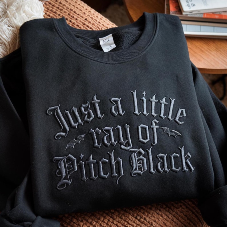 May include: Black crewneck sweatshirt with the embroidered phrase "Just a little ray of Pitch Black" in a gothic font. Two bat silhouettes are embroidered on the design. The sweatshirt is folded on a brown surface.