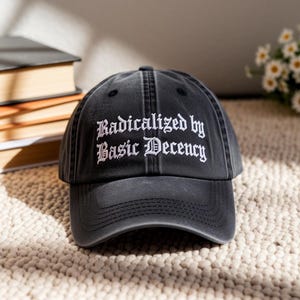 May include: Black baseball cap with the embroidered text "Radicalized by Basic Decency" in white gothic font. The cap is made of a soft, worn fabric and has a curved brim. The background includes a stack of books and a small bouquet of flowers.