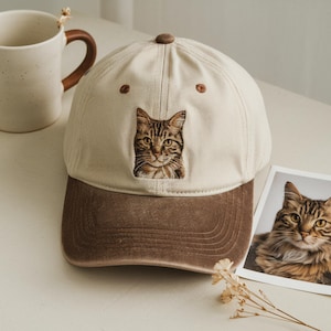 Peut inclure: Casquette de baseball beige avec une visière marron et un visage de chat brodé. La casquette a une visière incurvée et un bouton sur le dessus. Une photo de chat est à côté de la casquette.