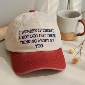 May include: A baseball cap with a cream-colored crown and a red brim. The cap has the embroidered text: "I WONDER IF THERE'S A HOT DOG OUT THERE THINKING ABOUT ME TOO". The cap has a red button on top.