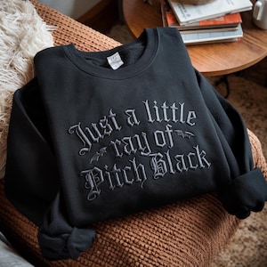 May include: Black sweatshirt with the embroidered phrase "Just a little ray of Pitch Black" in a gothic font. The sweatshirt is folded on a brown surface, with a cozy blanket and a small table in the background.