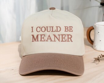 May include: A cream-colored baseball cap with a brown brim and a brown button on top. The front of the cap features the embroidered text "I COULD BE MEANER" in a brown, serif font. The cap has a structured crown and a curved visor.