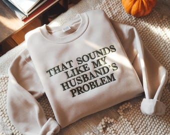 Może przedstawiać: Beżowa bluza z haftowanym napisem "THAT SOUNDS LIKE MY HUSBAND'S PROBLEM" w ciemnozielonej nici. Bluza jest złożona na teksturowanej powierzchni, z małą pomarańczową dynią w tle. Widoczne są książki.