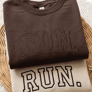 May include: A dark brown crewneck sweatshirt with the word RUN. embroidered in a matching dark brown thread, stacked on top of a cream-colored crewneck sweatshirt featuring the word RUN. embroidered in a bold black collegiate-style font.