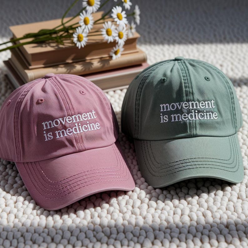 May include: Two baseball caps, one dusty rose and one sage green, are displayed. Each cap has the phrase "movement is medicine" embroidered in white. The caps are on a textured surface, with books and daisies in the background.