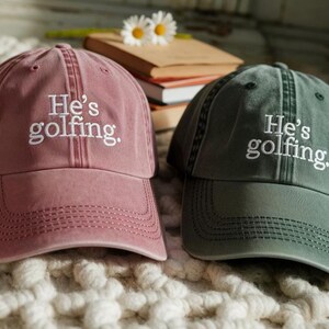 May include: Two baseball caps, one dusty rose and one dark green, are displayed. Both caps have the embroidered text "He's golfing." The caps are sitting on a textured, off-white surface.