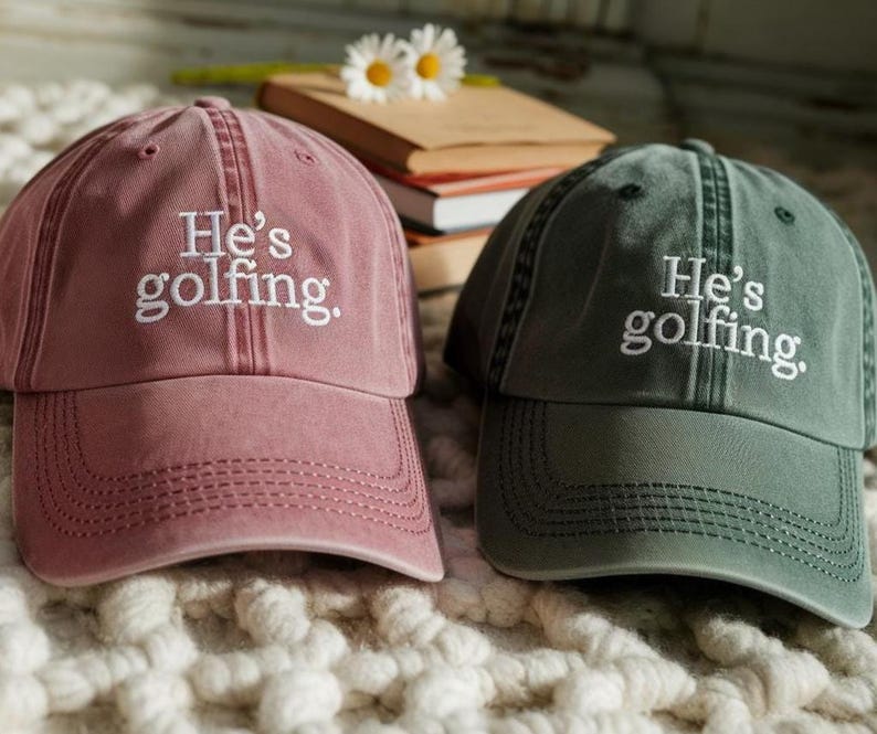 May include: Two baseball caps, one dusty rose and one dark green, are displayed. Both caps have the embroidered text "He's golfing." The caps are sitting on a textured, off-white surface.