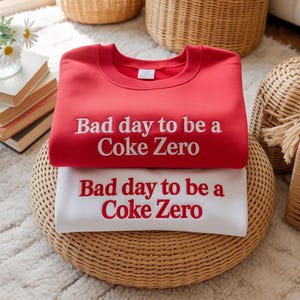 May include: Two sweatshirts, one red and one white, stacked on a woven surface. The red sweatshirt has white text that reads "Bad day to be a Coke Zero." The white sweatshirt has red text that reads the same. The sweatshirts are part of a clothing collection.