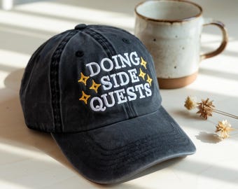 May include: A black baseball cap with the embroidered text "DOING SIDE QUESTS" in white and yellow. The cap is sitting next to a beige mug with a brown base and handle. Dried flowers are in the background.