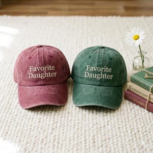May include: Two baseball caps, one burgundy and one green, are displayed. Each cap has the phrase "Favorite Daughter" embroidered in white. The caps are on a white, textured surface.