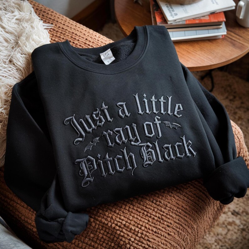 May include: Black sweatshirt with the embroidered phrase "Just a little ray of Pitch Black" in gray gothic lettering. The sweatshirt is folded on a brown surface, with a cozy blanket and a small table in the background.