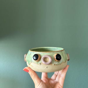 May include: A small, round ceramic bowl with a light beige base and a pale green glazed rim. The bowl features two large black circular eyes, a small pinkish-tan ring-shaped nose, and two small side protrusions resembling ears.