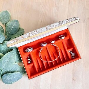May include: A rectangular jewelry box with a white lace-patterned lid and a vibrant orange velvet interior. The box contains several silver-toned rings, a thin twisted metal bracelet, and a pair of small stud earrings arranged in slots.