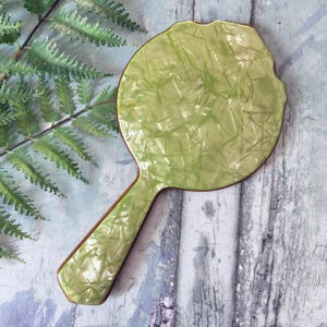 May include: A light green hand mirror with a textured surface and a brown border. The handle is also light green with a brown border. The mirror is on a weathered, light blue and gray wooden surface.