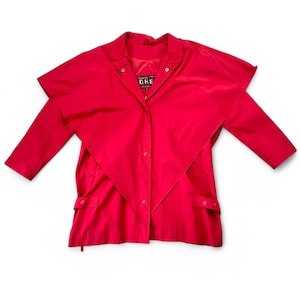 May include: A bright red jacket featuring a large, triangular cape-style overlay that extends over the shoulders and chest. The garment includes a front snap-button closure, long sleeves, and a black interior label with the text D.H.E. visible at the neck.