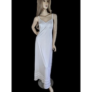 May include: A floor-length white slip dress featuring a V-neckline, thin shoulder straps, and a smooth, solid-colored fabric. The hem of the garment is finished with a decorative white lace trim detail that adds texture to the bottom edge.