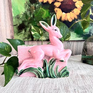 May include: A glossy pink ceramic deer figurine depicted in a leaping pose. The deer is positioned against a textured, light pink rectangular background with dark green painted grass at the base. The piece features delicate black painted eye details.