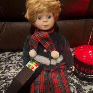 May include: A porcelain doll with blonde hair and blue eyes, wearing a dark green velvet jacket with white faux fur trim, a red and green plaid scarf with white pom-poms, and matching plaid trousers, holding a black electromagnetic field meter.