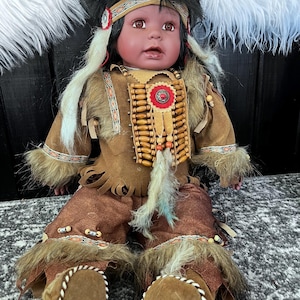 May include: A Native American-style doll with dark hair and a brown complexion. The doll wears a fringed suede outfit with bead and feather accents. The doll's outfit includes a headband, vest, and pants, and it is seated.