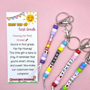 May include: Four personalized keychains with colorful plastic beads and letter cubes spelling names, attached to silver metal lobster clasps. Next to them is a rectangular card with a sun illustration and the text First Day of First Grade.