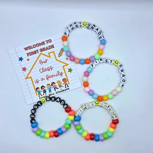 May include: Four circular bracelets made of colorful plastic beads and letter beads spelling FIRST GRADE with yellow smiley face beads. A white rectangular card features the text WELCOME TO FIRST GRADE, Our Class is a family, From: Mrs. Elizabeth.
