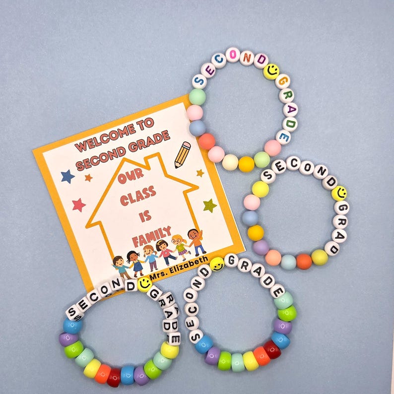 May include: Four colorful beaded bracelets spelling "SECOND GRADE" with smiley face beads. A welcome card with text "WELCOME TO SECOND GRADE" and "OUR CLASS IS FAMILY" is also in the image.