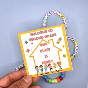 May include: A yellow square card with the text "WELCOME TO SECOND GRADE" and "OUR CLASS IS FAMILY" is held near a beaded bracelet. The card has a house graphic and illustrations of children. The bracelet spells "SECOND GRADE".