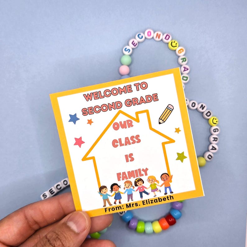 May include: A yellow square card with the text "WELCOME TO SECOND GRADE" and "OUR CLASS IS FAMILY" is held near a beaded bracelet. The card has a house graphic and illustrations of children. The bracelet spells "SECOND GRADE".