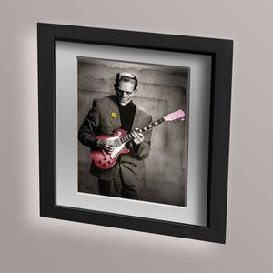 May include: A black and white photo of a man in a suit playing a red electric guitar with a pink pickguard. He is wearing a yellow smiley face pin on his jacket.