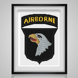 Op de afbeelding: Een zwart-witte kruissteekpatroon van de 101st Airborne Division patch. De patch heeft een witte adelaarskop met een rode snavel en gele ogen, en de woorden "AIRBORNE" in gele letters.