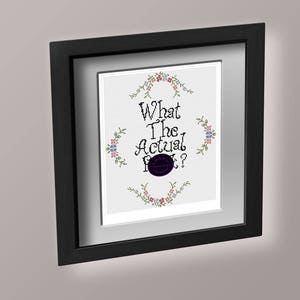 Pode incluir: Um bordado emoldurado com a frase "What the actual f***?" em letras pretas. O bordado é rodeado por uma borda de pequenas flores rosa e azuis.
