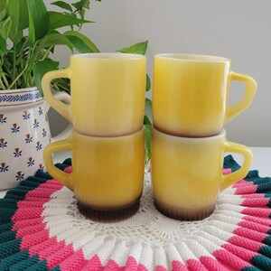 May include: Four yellow mugs stacked on a colorful, round, knitted coaster. The mugs have a classic design with a rounded body and a curved handle. A potted plant is in the background.