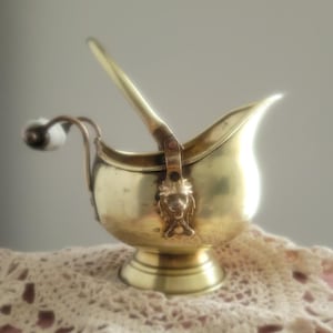 Peut inclure: Un sucrier vintage en laiton avec une poignée et un couvercle à charnière. Le bol présente une tête de lion décorative et une poignée blanche et noire. Le sucrier est posé sur un napperon en dentelle.