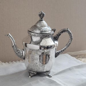 May include: A silver teapot with ornate floral and scrollwork detailing. The teapot features a curved spout, a decorative handle, and a lid with a finial. It stands on three small feet. The teapot is likely made of silver or a silver-plated metal.