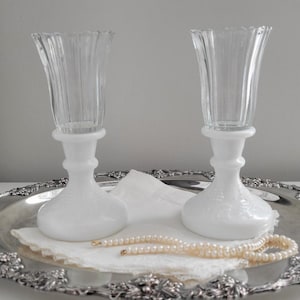 May include: Two clear glass flutes with a ribbed design, atop white milk glass bases. They sit on a silver-colored, ornate tray with a pearl necklace and a white doily. A classic, elegant display.