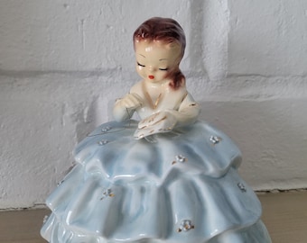 Peut inclure: Une figurine en porcelaine d'une jeune fille vêtue d'une robe bleue avec une bordure en dentelle blanche et des accents dorés. Elle tient un petit objet blanc dans ses mains et a les cheveux bruns.