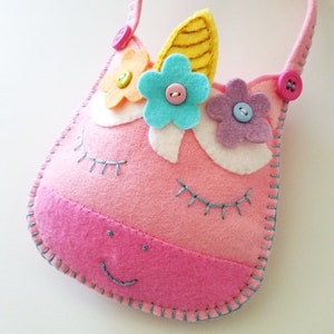 Puede incluir: Una bolsa de fieltro rosa en forma de unicornio con un cuerno amarillo, flores azules y rosas y detalles de botones. La bolsa tiene un asa y una cara sonriente.