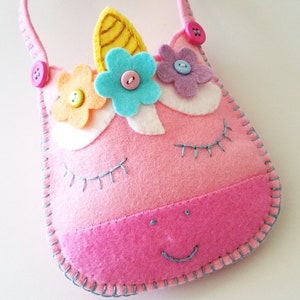 Puede incluir: Un bolso de unicornio de fieltro rosa con un cuerno amarillo, flores azules y rosas y botones rosas. El bolso tiene una sonrisa cosida azul y pesta&ntilde;as.