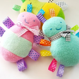 May include: Three colorful plush toys with ribbon tails. The toys are shaped like a bee, a turtle, and a pear. The bee is pink and green with a white and pink bow. The turtle is light blue with a gray and white bow. The pear is yellow and green with black and white polka dots.