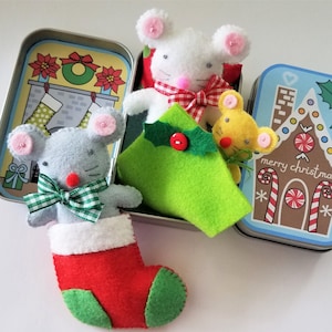 May include: A set of three felt mouse ornaments in a tin box. The mice are dressed in Christmas attire, including a stocking, a bow tie, and a green tree. The tin box has a gingerbread house design with the text "merry christmas".