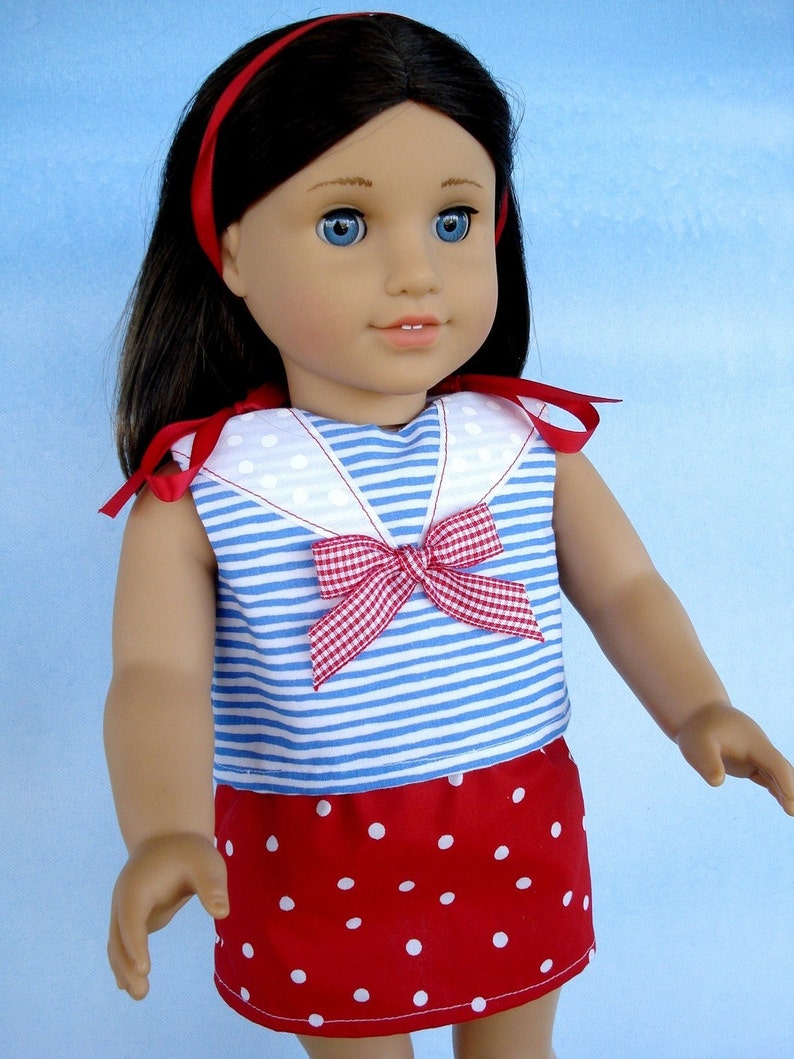 May include: A doll wearing a blue and white striped sailor top with a red and white gingham bow and a red skirt with white polka dots. The doll has long dark hair and blue eyes.