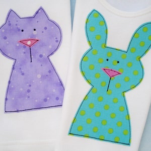 May include: Two white t-shirts with appliqued animal designs. One shirt features a purple cat with white polka dots. The other shirt features a blue bunny with green polka dots.
