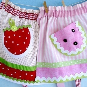 Puede incluir: Dos delantales de tela rosa y blanca con ribetes contrastantes. Un delantal tiene un bolsillo en forma de fresa con lunares blancos y un botón verde. El otro delantal tiene un bolsillo en forma de sandía con un borde festoneado verde y botones negros.