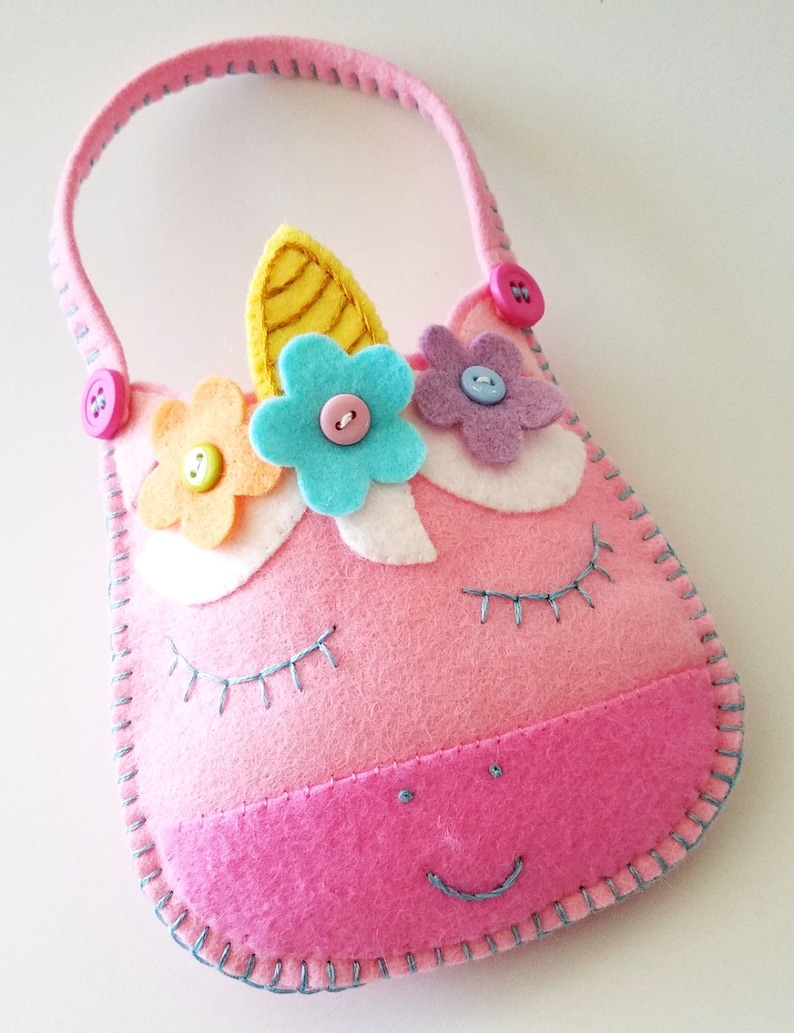 Puede incluir: Un bolso de unicornio de fieltro rosa con un cuerno amarillo, flores azules y rosas y botones rosas. El bolso tiene una sonrisa cosida azul y pesta&ntilde;as.
