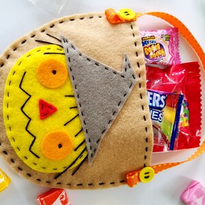 May include: A brown felt Halloween trick-or-treat bag shaped like a scarecrow with a yellow face, orange eyes, and a red nose. The bag has a handle made of orange ribbon with yellow polka dots and a yellow button. The bag is filled with sweets.