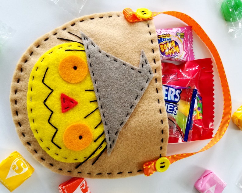 May include: A brown felt Halloween trick-or-treat bag shaped like a scarecrow with a yellow face, orange eyes, and a red nose. The bag has a handle made of orange ribbon with yellow polka dots and a yellow button. The bag is filled with sweets.